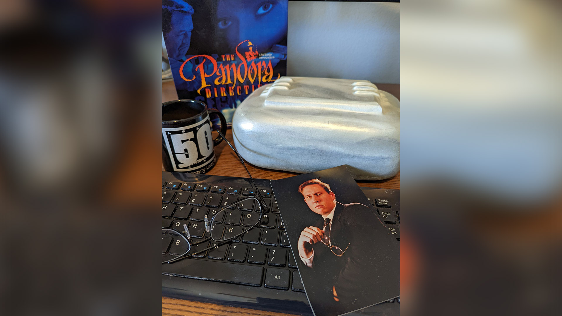 A photo of Mark Hulka from the Under a Killing Moon game boardroom sits on top of a computer keyboard. Around it are: a pair of glasses, a coffee mug, one of the Pandora Directive Puzzle box props, and an original Pandora Directive big box game box.